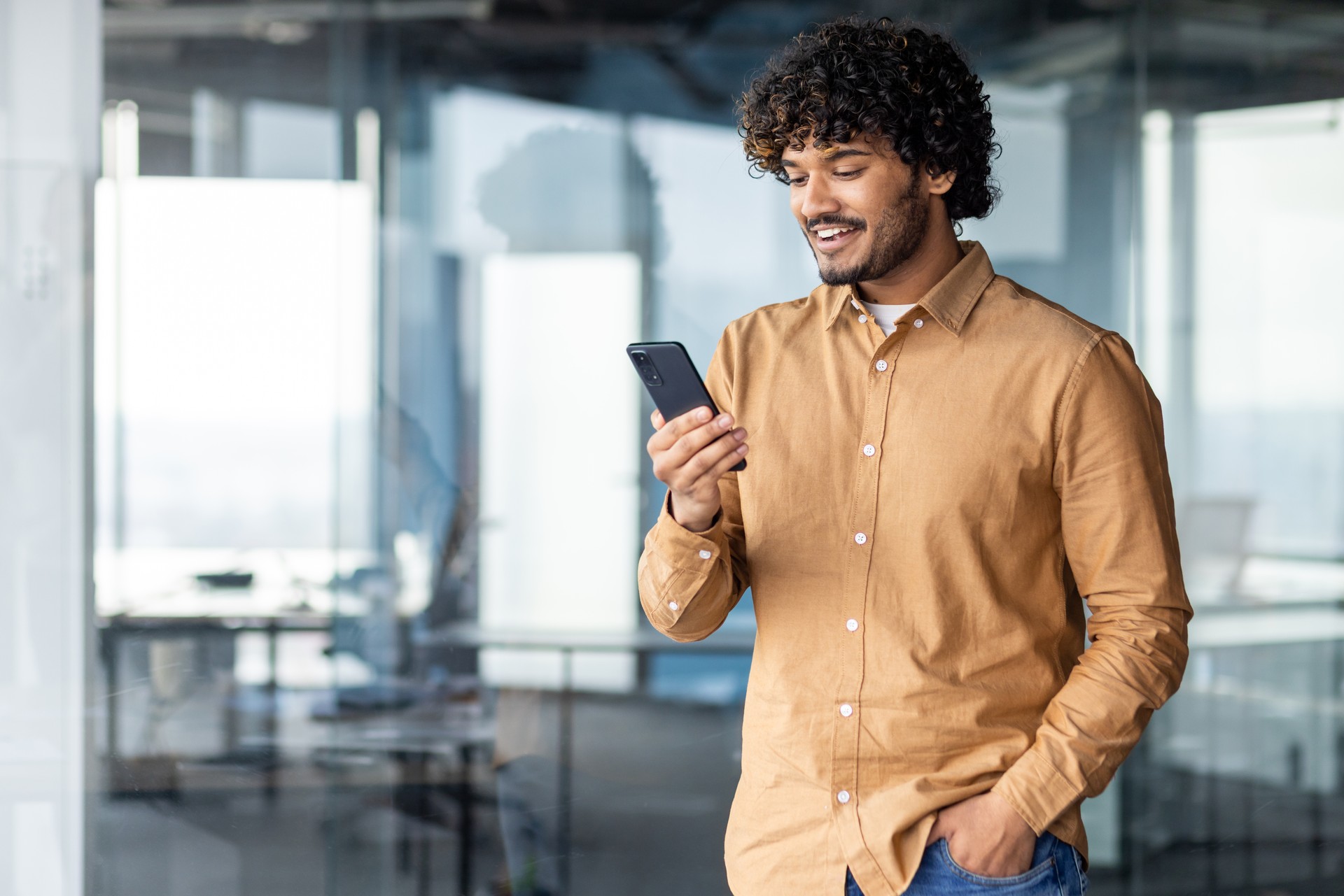 A young Hispanic stands inside office, the man holds the phone in his hands, the businessman browses online pages and uses applications, dials a message, makes a call, a smiling satisfied employee